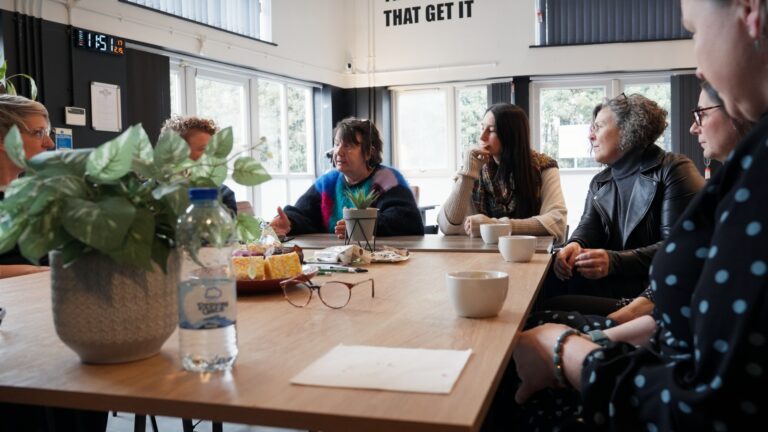 The photo is of a group of women sitting around a table talking in a workshop setting.