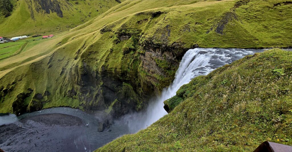 A photo of Skogafoss from the viewing platform above. White powerful water with bright green banks.