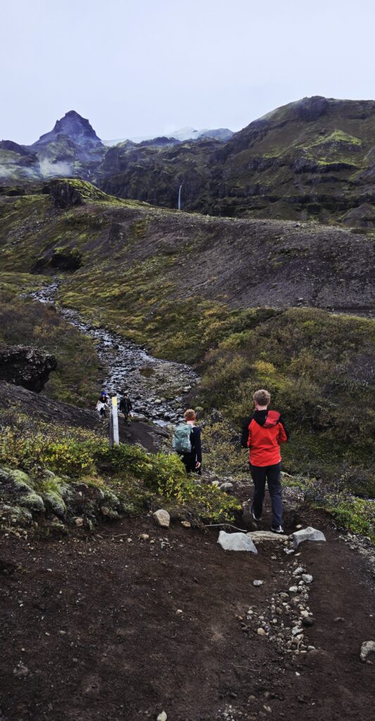 An image of walkers hiking up to The Mulagljufur Canyon. Grey and green in colour where moss is growing over the lava bed.