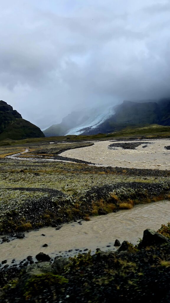 A iew from the south coast of a glacier making its way slowly down the mountain. In the foreground is the murky water, a mixture of glacier melt and rain,