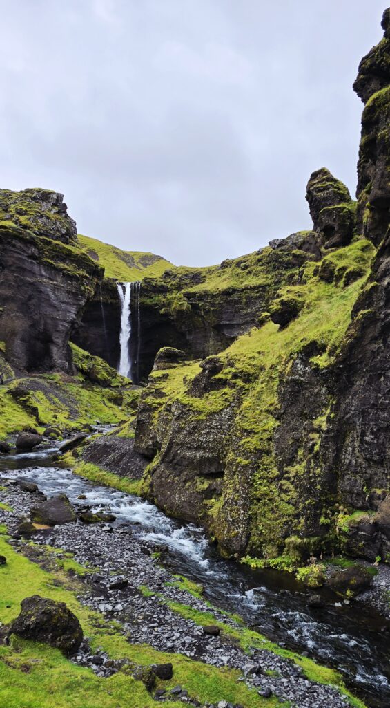 Kvernoss Waterfall - an photo of the walk up to the waterfall, showing the waterfall in the distance, black rock and lush green mossy sides, steeply meeting the river.