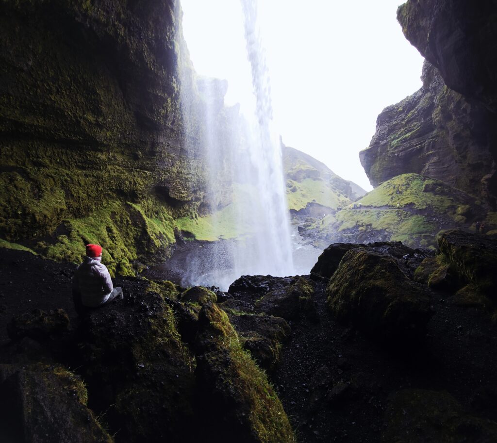 An image taken from behind the flow of water at The Kvernoss Waterfall. Dark black wet rocks, the powerful water falling and a green backdrop.