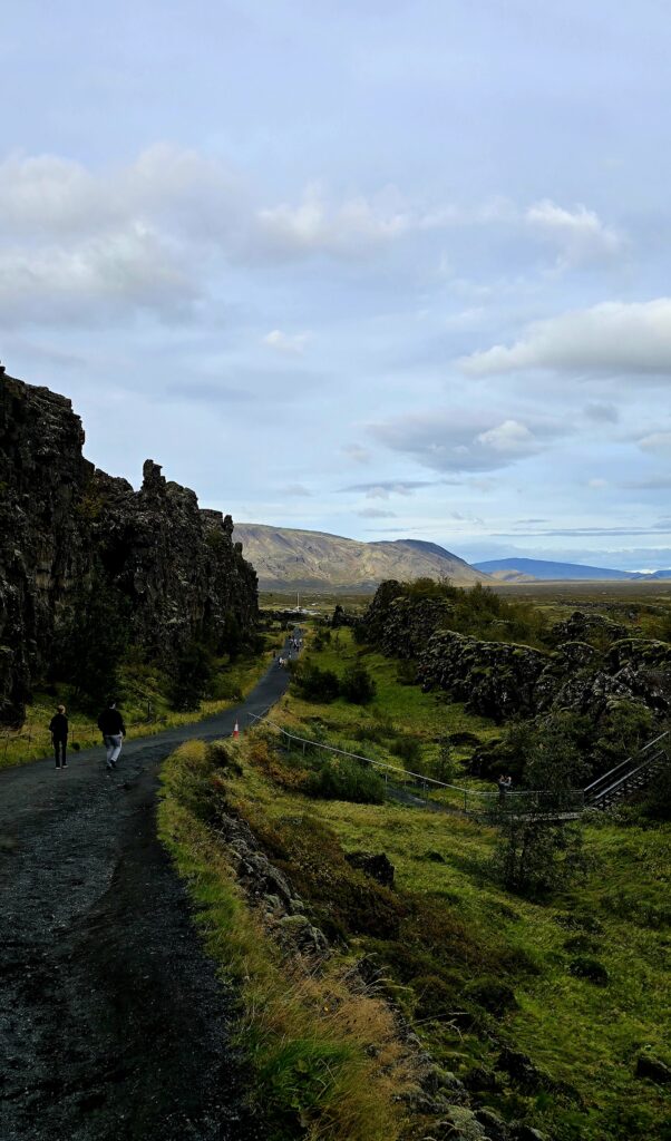 A landscape image that takes in the cliff of the falt lin, the path, green foliage and the hills in the distance at Thingvellir National Park.