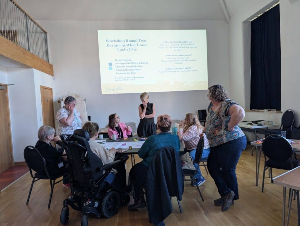 Bryony is standing, the rpesentation is on the wall behind her, while she listens to a member of the group giving feedback.
There are 8 people sat around the table. Phot credit Heather Birnie